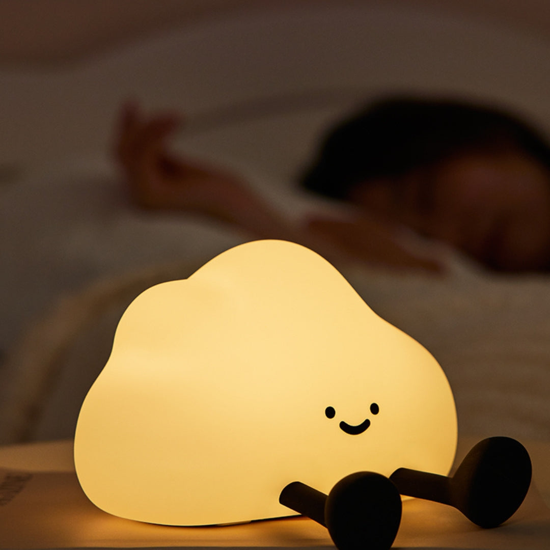 Cloud-shaped night light with a smiling face on a table, with a child lying in bed in the background.