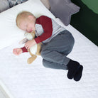 A young child sleeping on a white waterproof mattress protector designed for single beds, comfortably hugging a teddy bear.