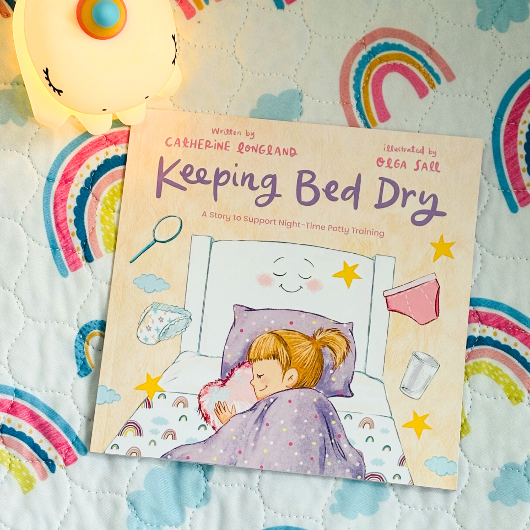 Children's book ' Keeping Bed Dry' with illustration of a child sleeping in bed with stars and a teddy bear, placed on a rainbow waterproof mattress protector.