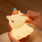 A night light turned on designed to look like a unicorn with a hand squashing the nose to show the silicone material.