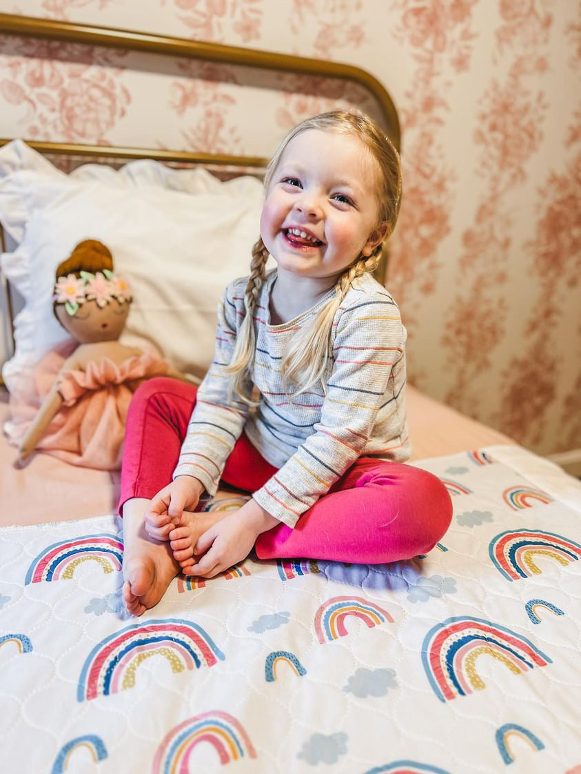Child sitting on a rainbow print Hygge waterproof bed pad on a pink bed